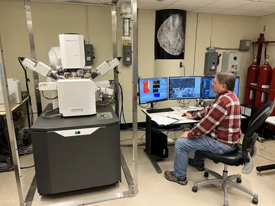 A man sits in front of a large, white machine and a row of computer screens in a lab setting.