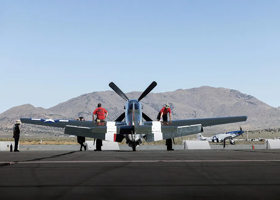 Speedball Alice, a restored dark green P-51D airplane with white stripes, is parked on a tarmac in the Reno desert, with mountains and blue sky in the background.