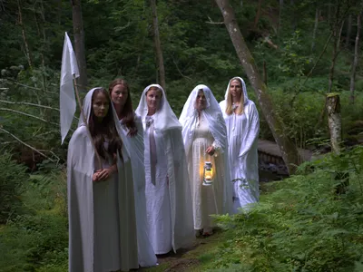 Five adults dressed in white dresses, robes, and veils pose along a forest path. One carries a lit lantern, and another carries a white flag.