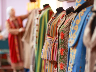 In the foreground, a lineup of mannequins each with colorful embroidered garments. At the end of the line, out of focus, a woman dresses a mannequin.