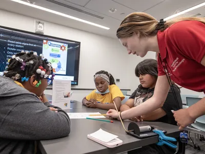 A young adult in a red polo points to a worksheet on a table. Three students are sitting at the table which has a table top rocket launcher, straw rocket, and table sign with instructions on top of it.