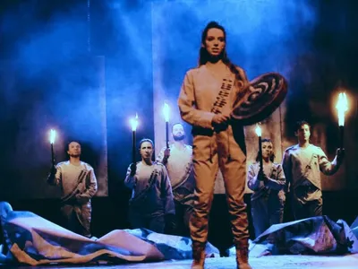 A woman stands on stage, holding a hand drum and a beater stick. Five actors behind her hold up torches. They all wearing matching white outfits with mock bandoliers. The stage is foggy and lit blue.