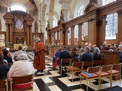 Interior of a church, with people seated in chairs and pews, and a person in reddish robe with black trim walking toward the pulpit.