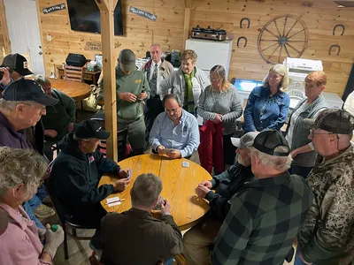 Four men sit around a round wooden table playing cards, while a crowd of others surround them.